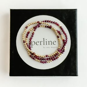 May include: Three beaded bracelets with gold-colored beads and dark red beads are displayed on a white circle inside a black box. The bracelets are stacked together. The text "perline by amy megan" is printed on the white circle.