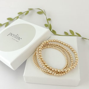 May include: A set of four gold beaded bracelets stacked on top of each other in a white box. The box has a label that says "perline by amy megan".