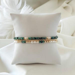 Puede incluir: Tres pulseras de cuentas sobre un cojín blanco. Las pulseras presentan cuentas verde esmeralda, pequeñas cuentas doradas y perlas blancas. Las pulseras están apiladas. El cojín está sobre tela blanca.