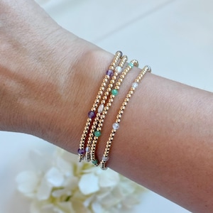 May include: Four gold beaded bracelets with colourful gemstones, including amethyst, emerald, and clear crystals. The bracelets are stacked on a wrist, showcasing the jewellery's design and colour variations. The bracelets are a delicate and elegant accessory.