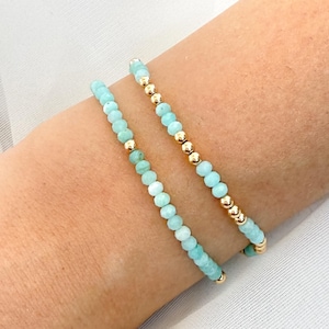 May include: Two beaded bracelets with light blue and gold beads. The bracelets are made of small, round, light blue beads interspersed with gold-colored beads. The bracelets are worn on a person's arm.