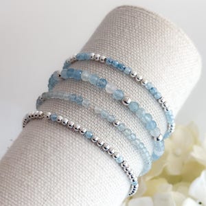 May include: A collection of delicate bracelets featuring light blue aquamarine beads and small silver beads. The bracelets are arranged around a cylindrical form. The aquamarine beads are of varying sizes, creating a layered effect.
