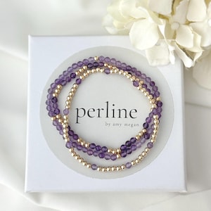 May include: Three delicate beaded bracelets arranged in a circular pattern on a white square box. The bracelets feature alternating small faceted purple amethyst beads and smooth metallic gold beads. The box displays the text perline by amy megan.