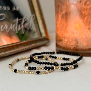 May include: Three stretch bracelets featuring a repeating pattern of small, round, metallic gold beads and faceted black beads. The bracelets are arranged in a cluster, showcasing the alternating texture of the smooth gold and the dark, angular black stones.