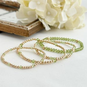 May include: Four gold beaded bracelets with small green gemstones. The bracelets are arranged on a white surface, with a blurred background of white flowers and a decorative frame.