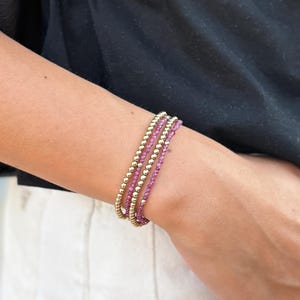 May include: A set of four bracelets. Two bracelets feature small, gold-colored beads, and the other two have small, pink beads. The bracelets are stacked on a wrist, with a black shirt and white pants in the background.