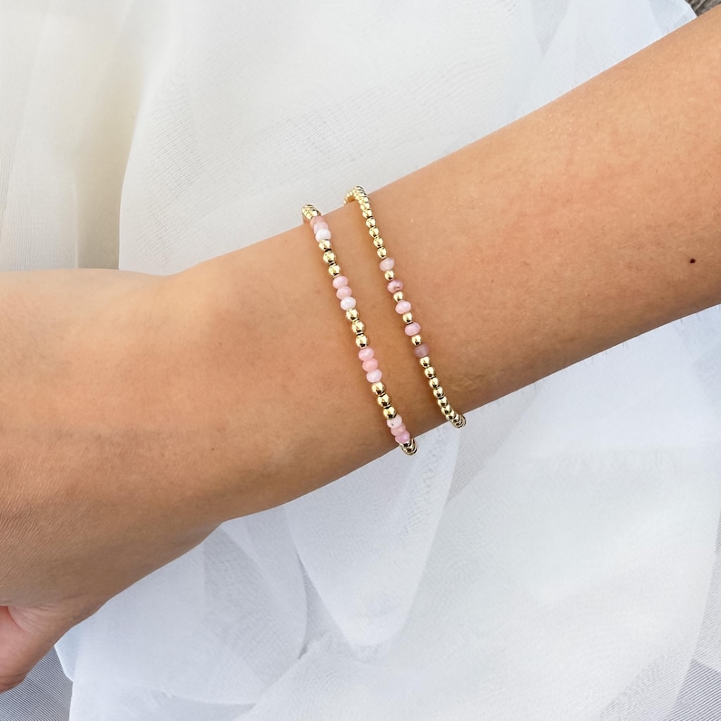 Puede incluir: Dos pulseras delicadas con peque&ntilde;as cuentas rosadas y blancas y detalles dorados. Las pulseras est&aacute;n apiladas en la mu&ntilde;eca de una persona.
