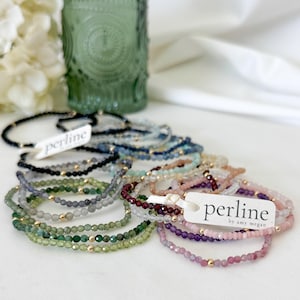 Puede incluir: Una colección de delicadas pulseras de cuentas en varios colores, incluyendo negro, verde, azul, morado y rosa. Cada pulsera presenta pequeñas cuentas facetadas y una cuenta dorada. Dos etiquetas blancas con el texto perline by amy megan están adjuntas.