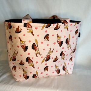 Puede incluir: Una bolsa de tela rosa con un asa negra y un patr&oacute;n repetido de un personaje de dibujos animados sosteniendo una pala. El personaje lleva una falda marr&oacute;n y tiene piel marr&oacute;n y cabello negro.