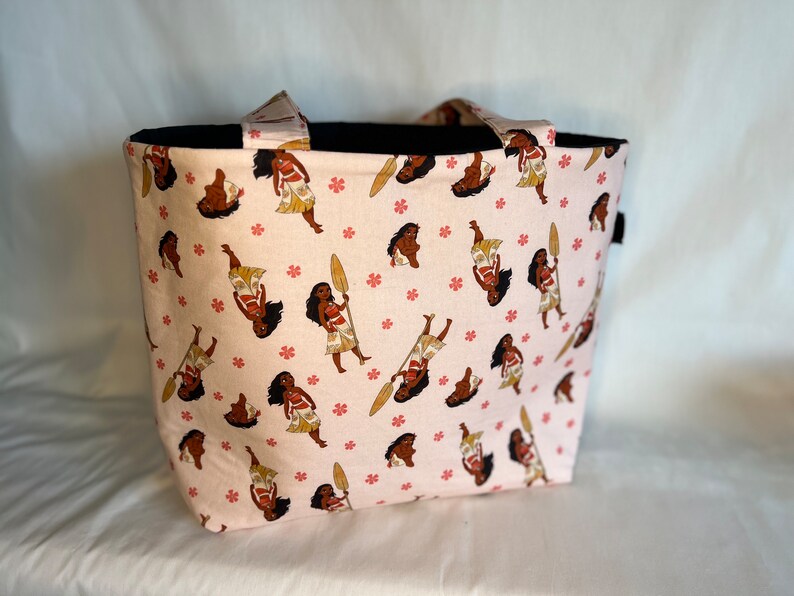 Puede incluir: Una bolsa de tela rosa con un asa negra y un patr&oacute;n repetido de un personaje de dibujos animados sosteniendo una pala. El personaje lleva una falda marr&oacute;n y tiene piel marr&oacute;n y cabello negro.