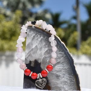 May include: A beaded bracelet featuring rose quartz and red beads, with a silver heart charm. The bracelet has a mix of light pink and white stones, with red beads and silver accents. The heart charm is intricately designed.