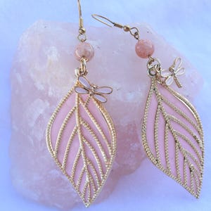 May include: A pair of dangle earrings featuring pink leaf-shaped pendants with gold-colored filigree and a small dragonfly charm. The earrings have a gold-colored hook and a pink bead at the top.