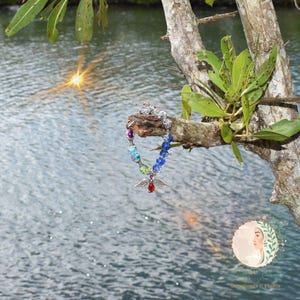 May include: A colorful beaded bracelet with a silver angel charm, hanging from a tree branch. The bracelet features a rainbow of beads, with a red gemstone at the charm's center. The background shows water and sunlight.