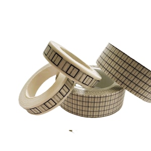 May include: Several rolls of off-white washi tape with a grid pattern in black. The tape is arranged in a dynamic composition against a white background, showcasing the product's design and texture.