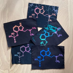 Könnte beinhalten: Fünf schwarze Stoffpatches mit jeweils einer darauf gedruckten chemischen Struktur. Die chemischen Strukturen sind in einem Farbverlauf von Pink, Blau und Lila gedruckt. Die chemischen Strukturen sind alle gleich, aber die Farben sind auf jedem Patch unterschiedlich.