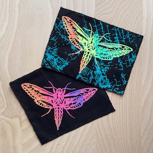 Könnte beinhalten: Zwei schwarze Stoffpatches mit einem regenbogenfarbenen Motten-Design. Die Motte ist auf einem schwarzen Hintergrund mit einem blauen und weißen Muster gedruckt.