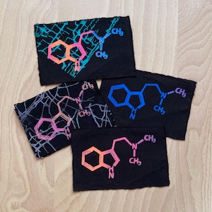 Könnte beinhalten: Vier schwarze Stoffflicken mit farbenfrohen Molekularstruktur-Designs. Die Designs sind in Blau-, Rosa-, Orange- und Weißtönen gehalten. Die Flicken sind auf einer hellen Holzoberfläche angeordnet.