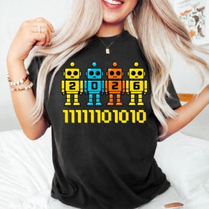 Peut inclure: T-shirt noir avec quatre robots colorés affichant les chiffres "2026" et un motif de code binaire. Les robots sont jaunes, bleus et orange, avec du texte jaune.