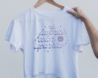 Puede incluir: Camiseta blanca recortada con un gráfico rosa y morado que dice "mi color favorito es brillos" con estrellas alrededor del texto.