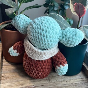 Puede incluir: Un juguete de peluche tejido a crochet con una cabeza azul claro y un cuerpo marr&oacute;n. El juguete tiene una bufanda blanca de crochet alrededor del cuello.