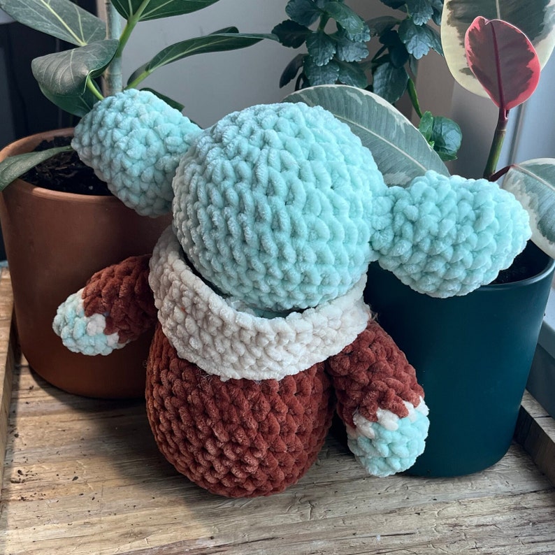 Puede incluir: Un juguete de peluche tejido a crochet con una cabeza azul claro y un cuerpo marr&oacute;n. El juguete tiene una bufanda blanca de crochet alrededor del cuello.