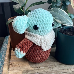 Puede incluir: Un juguete de peluche tejido a crochet con forma de personaje con ojos grandes y redondos. El juguete est&aacute; hecho de hilo azul claro y marr&oacute;n y tiene una bufanda de crochet blanca.
