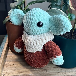 Puede incluir: Un juguete de peluche tejido a crochet con forma de personaje con orejas grandes, cuerpo marr&oacute;n y una bufanda blanca. El juguete tiene una cabeza azul claro con un ojo negro.
