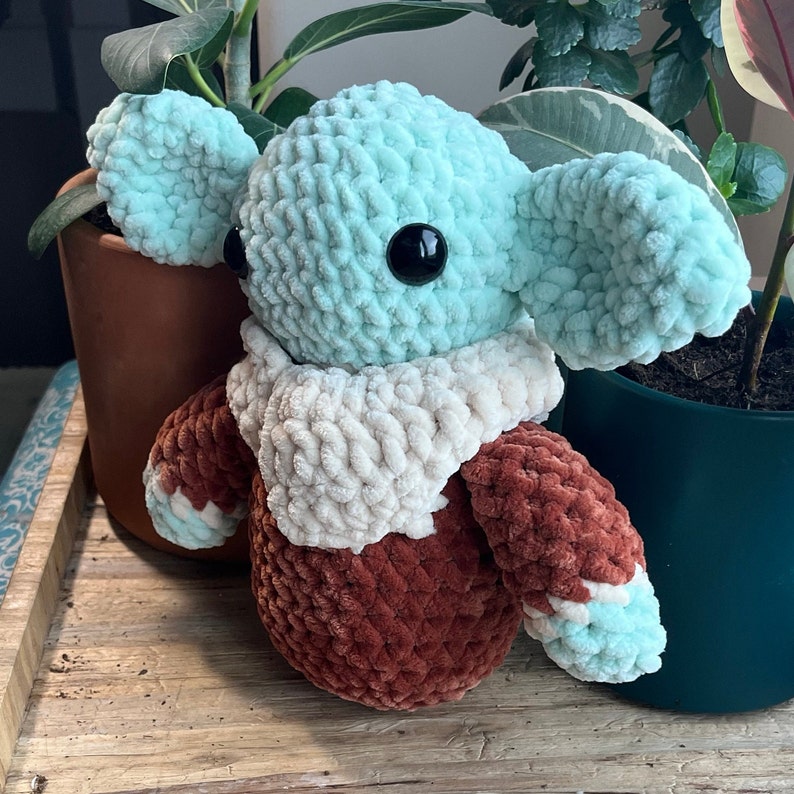 Puede incluir: Un juguete de peluche tejido a crochet con forma de personaje con orejas grandes, cuerpo marr&oacute;n y una bufanda blanca. El juguete tiene una cabeza azul claro con un ojo negro.