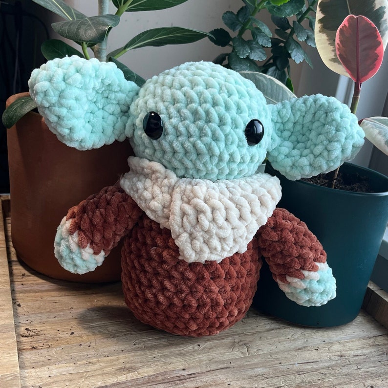 Puede incluir: Un juguete de peluche tejido a crochet con forma de un personaje de la franquicia Star Wars. El juguete es verde y marr&oacute;n con una bufanda blanca.