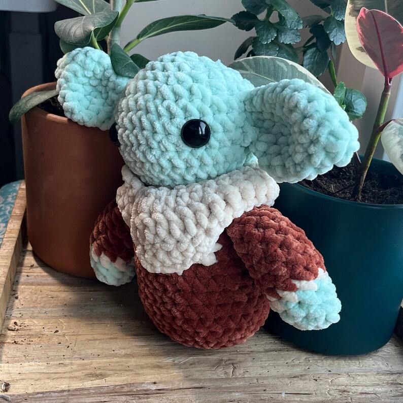 Puede incluir: Un juguete de peluche tejido a crochet con forma de personaje de la franquicia Star Wars. El juguete es de color verde claro con detalles marrones y blancos. El juguete est&aacute; sentado sobre una superficie de madera.
