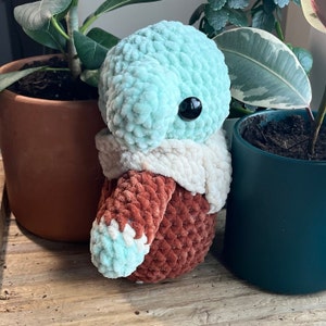 Puede incluir: Un juguete de peluche tejido a crochet con una cabeza azul claro, un cuerpo marr&oacute;n y una bufanda blanca. El juguete tiene un ojo negro grande y est&aacute; sentado sobre una superficie de madera.