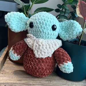 Puede incluir: Un juguete de peluche tejido a crochet con forma de personaje de la franquicia Star Wars. El juguete es verde, marr&oacute;n y blanco y tiene ojos grandes.