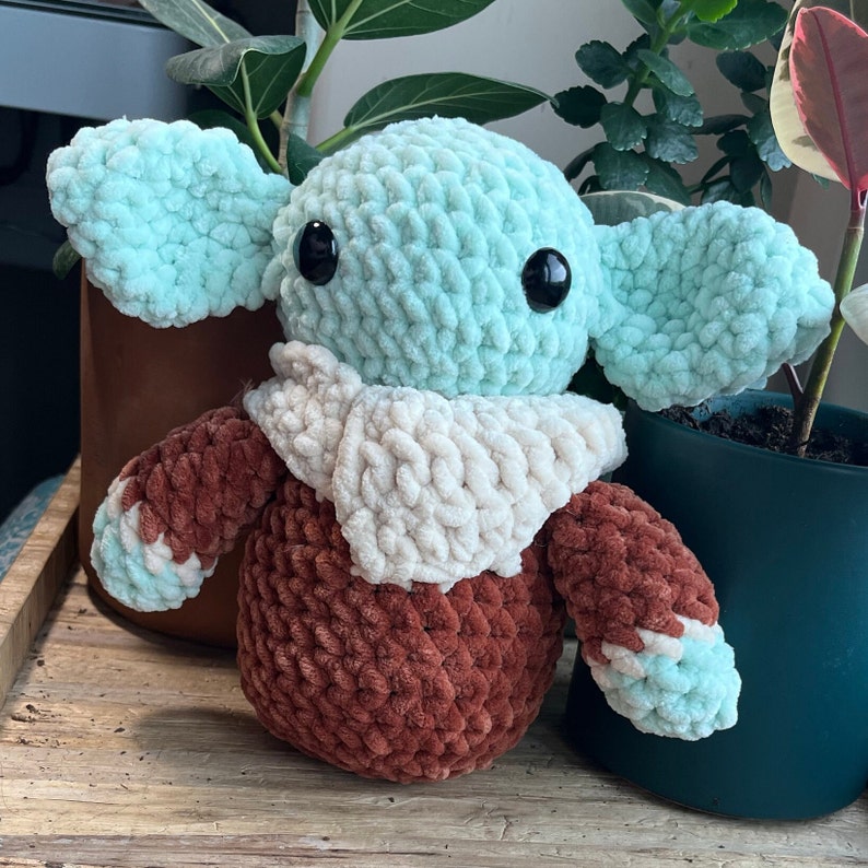 Puede incluir: Un juguete de peluche tejido a crochet con forma de personaje de la franquicia Star Wars. El juguete es verde, marr&oacute;n y blanco y tiene ojos grandes.