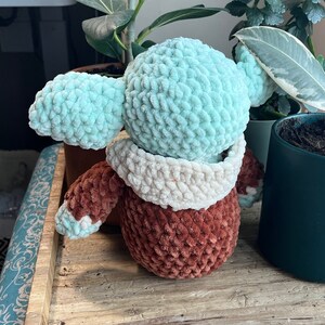 Puede incluir: Un juguete de peluche tejido a crochet con forma de personaje con cabeza azul claro, cuello blanco tejido a crochet y cuerpo marr&oacute;n.