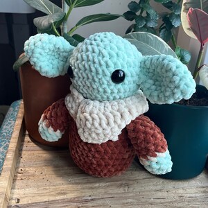 Puede incluir: Un juguete de peluche tejido a crochet con forma de personaje con orejas grandes, cuerpo marr&oacute;n y una bufanda blanca. El juguete tiene ojos grandes y negros y una cabeza verde claro.
