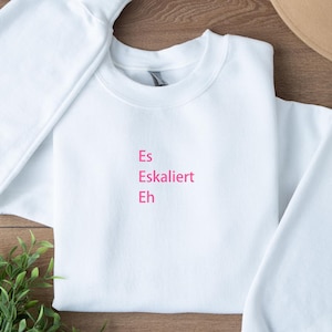 Könnte beinhalten: Ein weißes Sweatshirt mit Rundhalsausschnitt und dem pinkfarbenen Text "Es Eskaliert Eh". Das Sweatshirt liegt auf einer Holzoberfläche, mit einem beigen Hut und einer grünen Pflanze im Hintergrund.
