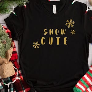 May include: Black crew neck t-shirt with the words "SNOW CUTE" in gold lettering, along with gold snowflake graphics. The shirt is displayed with Christmas decorations, including socks, lights, and a small tree.