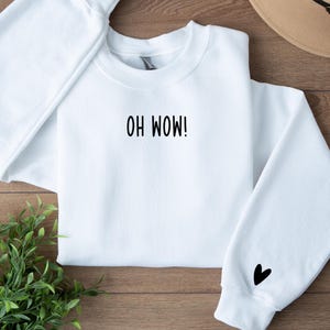 Peut inclure: Sweat-shirt blanc avec le texte "OH WOW!" imprimé sur le devant et un petit cœur noir sur la manche gauche.