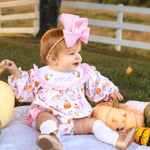May include: A baby wearing a pink bow headband, a white and pink pumpkin-themed outfit, and gold shoes sits on a white blanket. The outfit has the word "one" embroidered on the collar. Pumpkins surround the baby.
