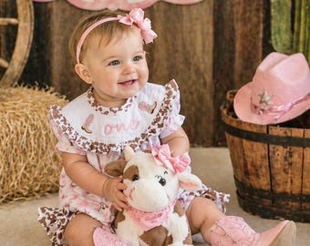 Peut inclure: Un bébé vêtu d'une tenue sur le thème cow-girl, comprenant une robe rose, des bottes et un bandeau, sourit en tenant une vache en peluche. L'arrière-plan comprend une pancarte avec le mot "ONE" et un chapeau de cow-boy rose.