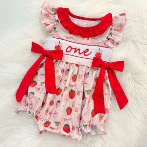 May include: A pink and red baby onesie with a strawberry print and the word "one" embroidered on the front. The onesie has red bows on the sides and a ruffled neckline.