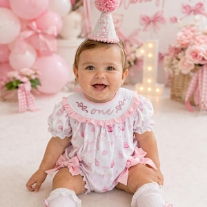 May include: A baby wearing a pink and white outfit with a matching party hat. The outfit has a cupcake and bow pattern. The hat has a pink pom-pom on top and the number "1". The background includes pink balloons and a sign that says "Girls Just Wanna Have Fun".