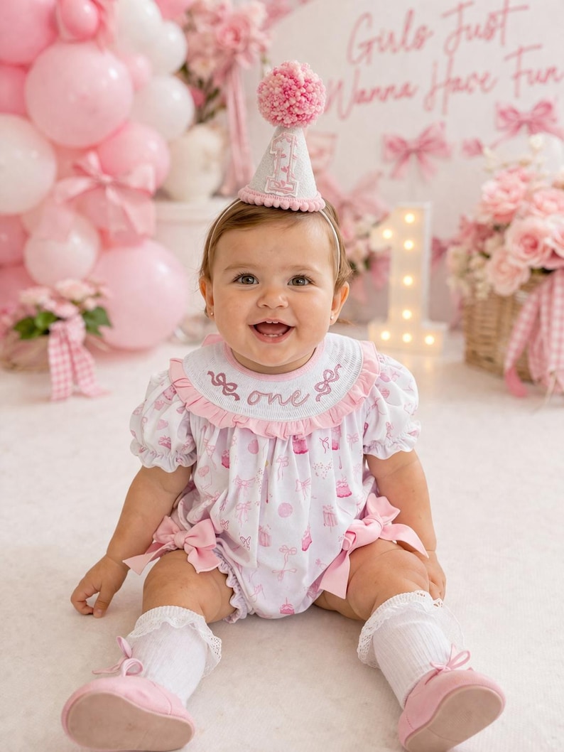 May include: A baby wearing a pink and white outfit with a matching party hat. The outfit has a cupcake and bow pattern. The hat has a pink pom-pom on top and the number "1". The background includes pink balloons and a sign that says "Girls Just Wanna Have Fun".