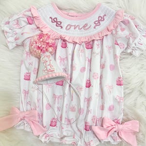 May include: A white and pink baby romper with a matching party hat. The romper features a pattern of bows, cakes, and balloons. The collar is embroidered with the word "one". Large pink bows are attached at the waist. The party hat is decorated with a pink pom-pom.