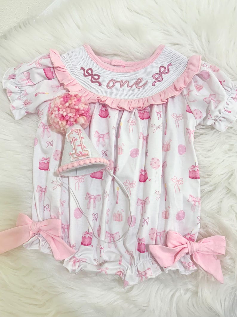 May include: A white and pink baby romper with a matching party hat. The romper features a pattern of bows, cakes, and balloons. The collar is embroidered with the word "one". Large pink bows are attached at the waist. The party hat is decorated with a pink pom-pom.