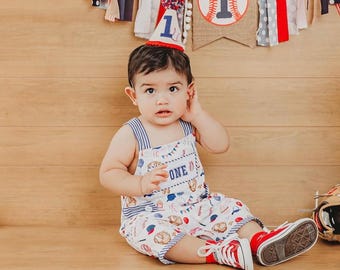 May include: A baby wearing a baseball-themed outfit, including a bib overall with the word "ONE" and a baseball print, a party hat, and red sneakers. A baseball and a baseball glove are nearby. A banner with baseball imagery hangs in the background.