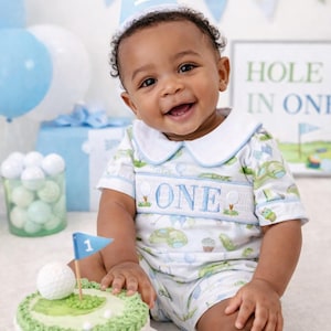 May include: A short-sleeved white romper with a blue collar and a smocked chest panel featuring the word ONE and golf-themed embroidery. The romper is paired with a light blue party hat with a white pom-pom and the number 1 on the front.