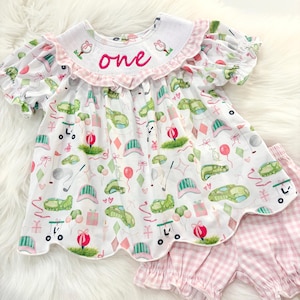 May include: A white baby outfit with a golf-themed print, including golf carts, hats, and balloons. The outfit features a ruffled collar with the word "one" embroidered in pink. Matching pink gingham shorts complete the set.