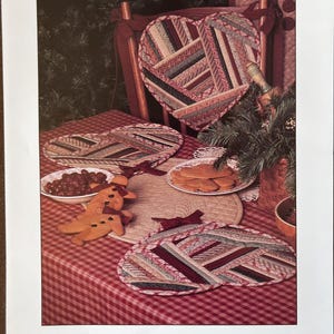 May include: A printed pattern titled "Holiday Hearts" featuring heart-shaped quilted placemats and a table setting with cookies and berries. The placemats have a patchwork design in shades of red, beige, and green.
