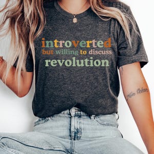 May include: A dark heather grey t-shirt with the text "introverted but willing to discuss revolution" printed in colorful letters.